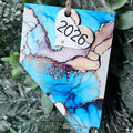 A decorative hanging ornament shaped like the state of Nevada, featuring a blue, beige, and white marbled pattern with gold veining. A small white heart tag is attached and reads “2026,” suspended by a twine string against a background of green pine branches.