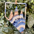 A decorative hanging ornament shaped like the state of Nevada, featuring a cutout landscape design with pine trees and blue, beige, and brown agate-like stone patterns, suspended by a twine string against green foliage.