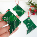 Hand holding a green circuit board Christmas tree ornament with more ornaments and holly in the background.