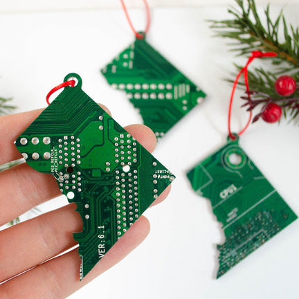 Hand holding a green circuit board Christmas tree ornament with more ornaments and holly in the background.