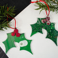 Green holly ornament made from recycled computer circuit board with red berries on a white surface with Christmas tree branches.