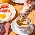 Person pouring a can of coffee into a cup with breakfast items on a table.