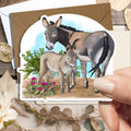 A hand holds a die-cut greeting card featuring an illustration of a donkey and foal, with a brown envelope behind it. The card is photographed on a rustic wooden surface with dried flowers, leaves, and small decorative props.