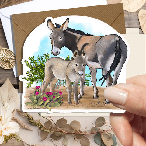 A hand holds a die-cut greeting card featuring an illustration of a donkey and foal, with a brown envelope behind it. The card is photographed on a rustic wooden surface with dried flowers, leaves, and small decorative props.