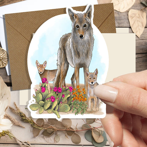 A hand holds a die-cut sticker featuring an illustrated coyote family with cacti and flowers, set against a rustic flat-lay background with dried flowers and envelopes.