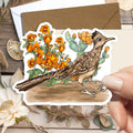 A hand holds a die-cut sticker featuring an illustrated brown bird standing among orange flowers and green cacti, placed over a styled flat-lay with a kraft envelope, dried leaves, and a white flower on a rustic wood surface.