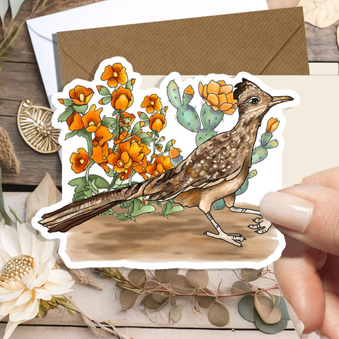 A hand holds a die-cut sticker featuring an illustrated brown bird standing among orange flowers and green cacti, placed over a styled flat-lay with a kraft envelope, dried leaves, and a white flower on a rustic wood surface.