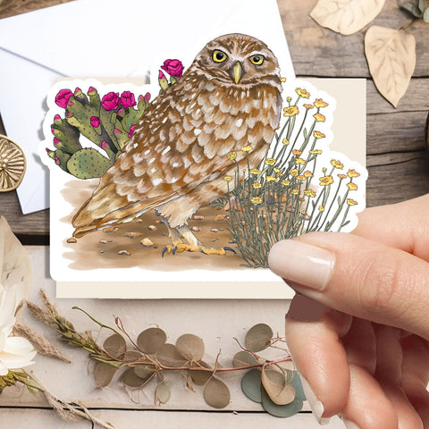 A hand holds a die-cut sticker featuring a detailed watercolor illustration of a brown owl with cactus and wildflowers, displayed on a rustic wooden surface with dried botanicals and an envelope.