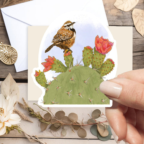 A hand holds a die-cut art card featuring a brown sparrow perched on a green prickly pear cactus with pink flowers, set against a rustic flat-lay background with dried flowers and leaves.