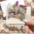 A hand holds a die-cut sticker featuring a painted tortoise surrounded by flowers, placed over a cream envelope on a styled flat-lay with dried leaves, a white flower, and a wooden surface.