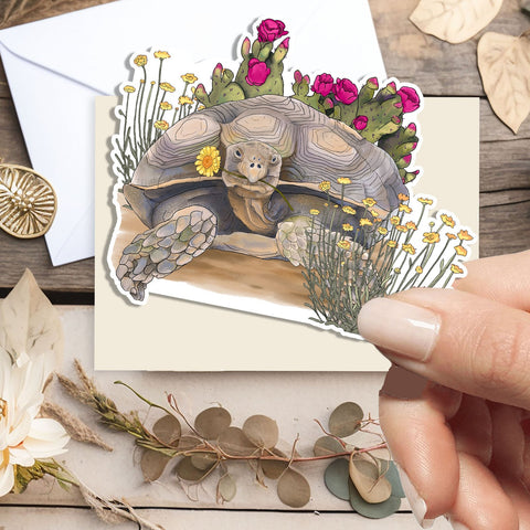 A hand holds a die-cut sticker featuring a painted tortoise surrounded by flowers, placed over a cream envelope on a styled flat-lay with dried leaves, a white flower, and a wooden surface.