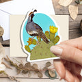 A hand holds a die-cut sticker featuring a desert quail perched on a prickly pear cactus, set against a light blue sky and yellow flowers. The sticker is displayed on a beige card over a rustic flat-lay background with dried flowers, a white envelope, and decorative leaves.