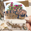 A hand holds a die-cut art card featuring a painted gila monster with cactus flowers, styled on a rustic wood background with dried flowers and leaves.