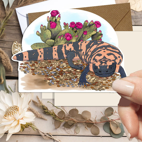 A hand holds a die-cut art card featuring a painted gila monster with cactus flowers, styled on a rustic wood background with dried flowers and leaves.