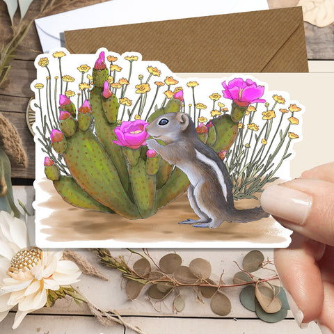 A hand holds a die-cut sticker featuring an illustrated chipmunk beside a green cactus with bright pink flowers, set against a styled flat-lay background with dried leaves, a white flower, and a kraft envelope.