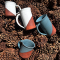 Four ceramic mugs with geometric designs on a bed of pine cones.