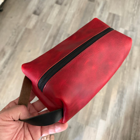 Red leather pouch with black zipper held by a hand on a wooden floor background