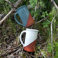 Two ceramic mugs, one blue and one white with a red base, are placed on a bed of leaves and twigs.