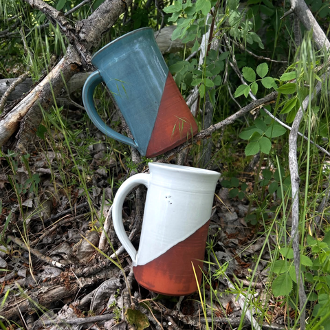 Two ceramic mugs, one blue and one white with a red base, are placed on a bed of leaves and twigs.