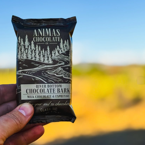 Hand holding a package of Animas Chocolate River Bottom Chocolate Bark against a blurred natural background.