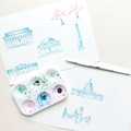 Artistic illustrations of buildings on paper with a watercolor palette and brush.
