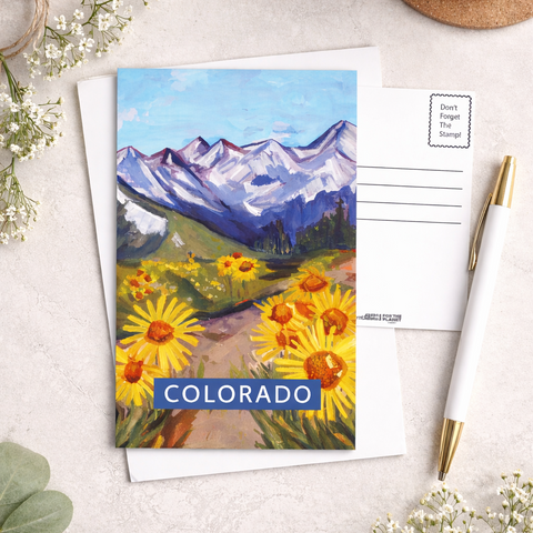 Postcard with a mountain and flower design labeled 'Colorado' on a light surface with a cup of coffee and decorative elements.