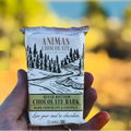 Hand holding a bar of Animas Chocolate Co. River Bottom Chocolate Bark with a scenic design on the packaging.