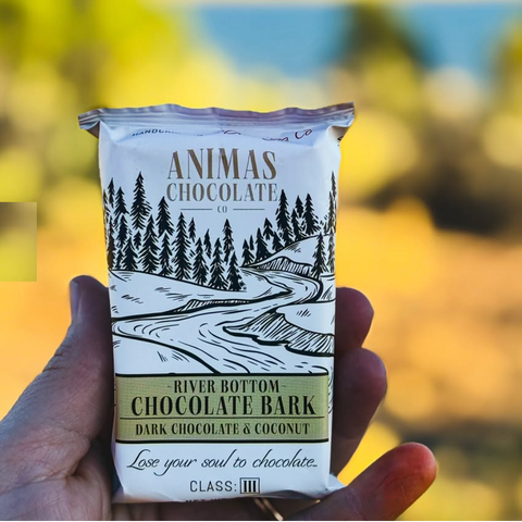 Hand holding a bar of Animas Chocolate Co. River Bottom Chocolate Bark with a scenic design on the packaging.